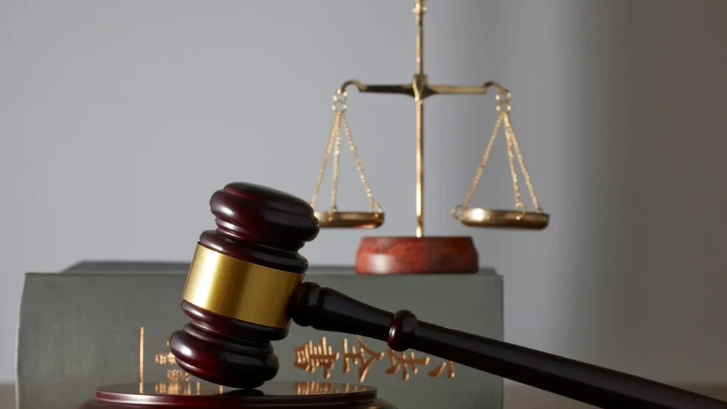 A wooden judge's gavel resting on a sound block in the foreground, with the scales of justice and legal law books blurred in the background, symbolizing the legal proceedings for robbery charges and sentencing.
