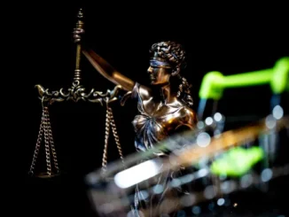 A bronze statue of Lady Justice holding scales, positioned behind a blurred metal shopping cart against a black background.