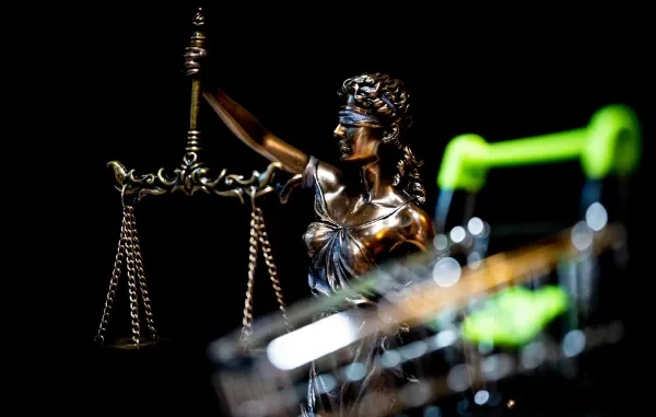 A bronze statue of Lady Justice holding scales, positioned behind a blurred metal shopping cart against a black background.