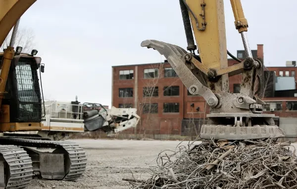 A heavy-duty excavator using a large industrial circular electromagnet to lift and sort a pile of twisted metal scrap at a construction site.