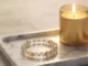 A handmade custom bracelet featuring clear and amber-toned faceted gemstone beads, resting on a white marble tray next to a glowing gold candle.