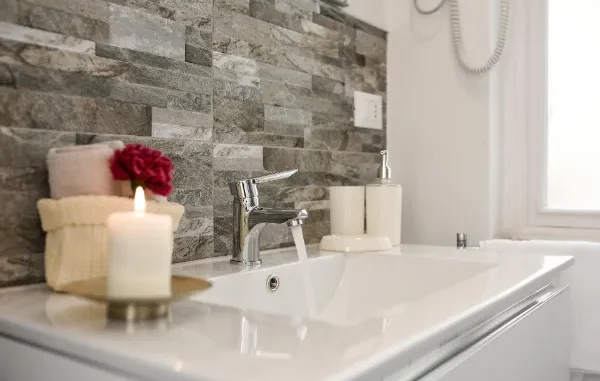 A sparkling clean white bathroom sink with a chrome faucet running water, featuring a textured stone tile backsplash and organized toiletries.