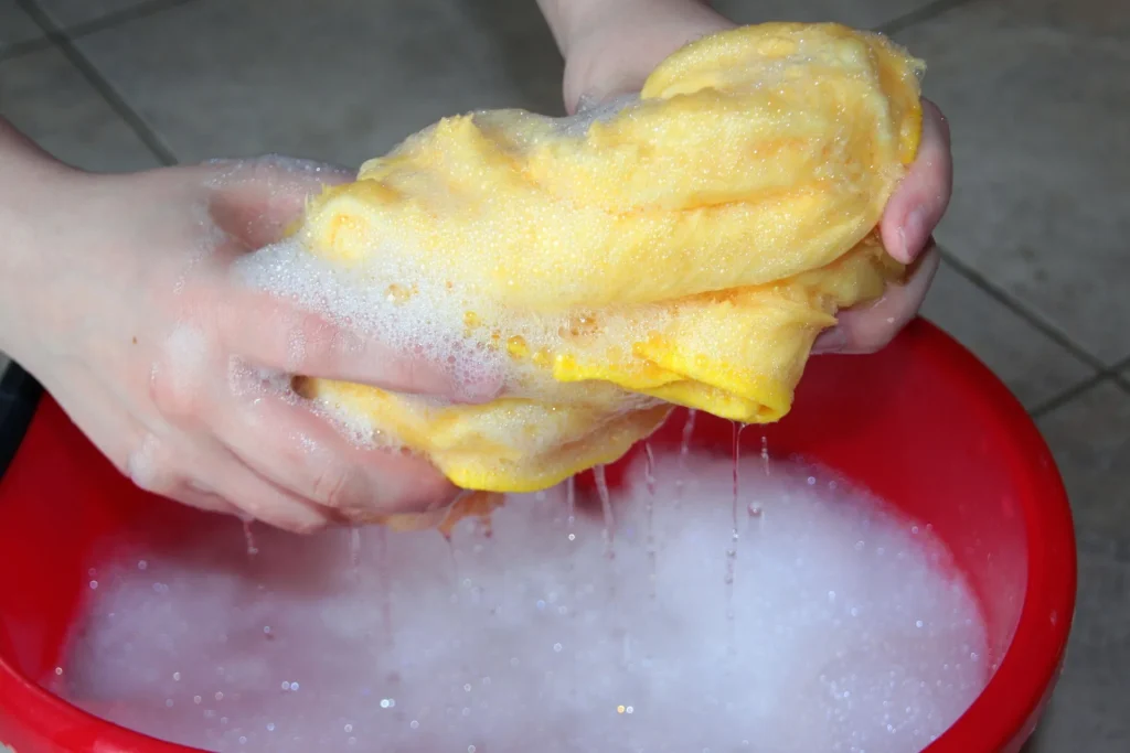 Close-up of hands wringing out a yellow microfiber cloth over a red bucket filled with soapy foam, demonstrating deep cleaning and sanitization techniques.