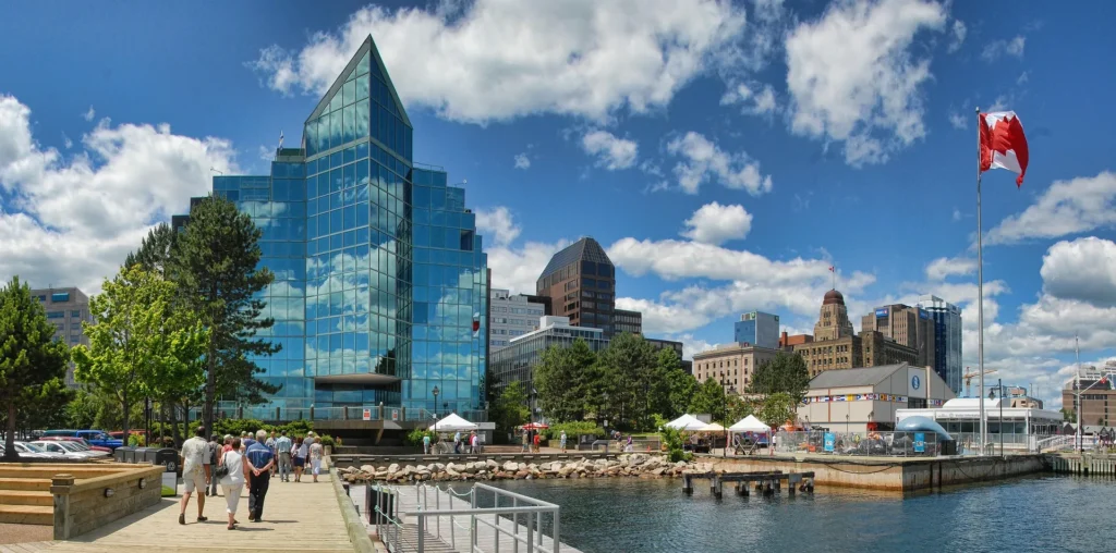 A vibrant view of the Halifax waterfront in Nova Scotia, Canada, featuring modern glass office buildings, a bustling boardwalk with people, and the Canadian flag, representing a thriving business environment for entrepreneurs.