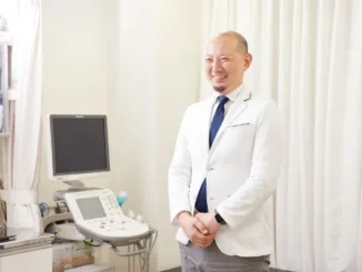 A smiling male medical professional in a white lab coat standing in a clinical setting next to a Toshiba ultrasound machine with a display monitor and control panel.