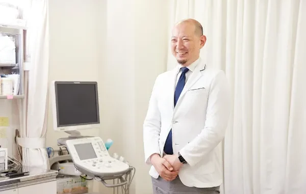 A smiling male medical professional in a white lab coat standing in a clinical setting next to a Toshiba ultrasound machine with a display monitor and control panel.