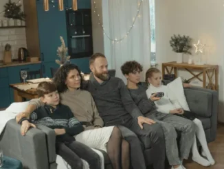 A family of five sits together on a couch in a cozy, lit living room watching television, illustrating household energy usage.