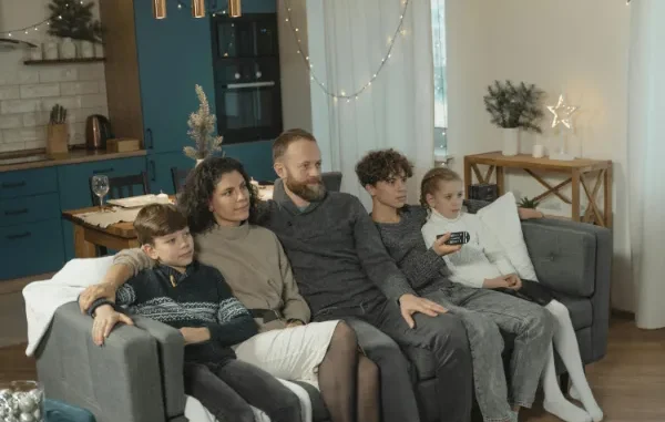 A family of five sits together on a couch in a cozy, lit living room watching television, illustrating household energy usage.