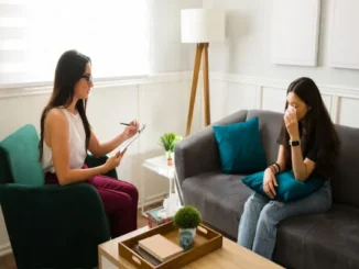 A female therapist in a bright, modern office taking notes on a clipboard while a young woman sitting on a gray sofa wipes her eyes with a tissue, illustrating a supportive mental health treatment session.