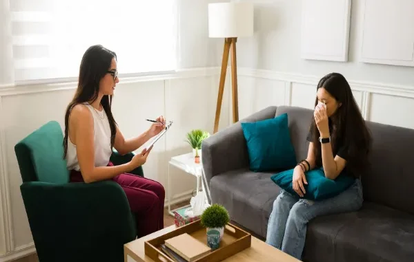A female therapist in a bright, modern office taking notes on a clipboard while a young woman sitting on a gray sofa wipes her eyes with a tissue, illustrating a supportive mental health treatment session.