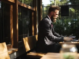 A man with a beard wearing a charcoal suit sits at an outdoor wooden table, focused on his laptop while researching intellectual property law.
