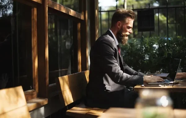 A man with a beard wearing a charcoal suit sits at an outdoor wooden table, focused on his laptop while researching intellectual property law.