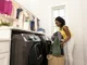 A woman in a bright laundry room loading green and purple clothes into a modern, dark stainless steel front-load washing machine.