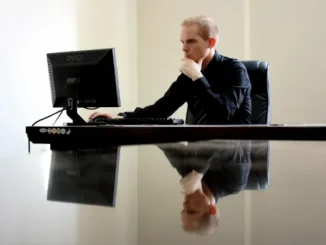 A professional man in a black shirt sitting at a desk, looking thoughtfully at a computer monitor. His reflection is visible on the polished desk surface.