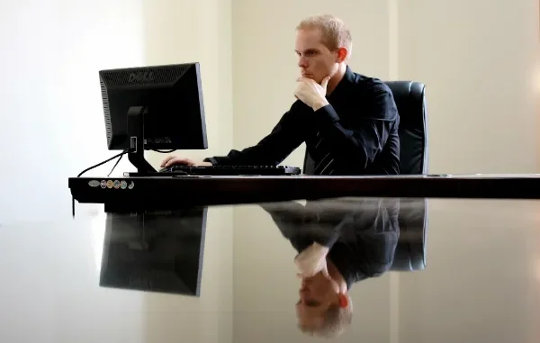 A professional man in a black shirt sitting at a desk, looking thoughtfully at a computer monitor. His reflection is visible on the polished desk surface.