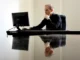 A professional man in a black shirt sitting at a desk, looking thoughtfully at a computer monitor. His reflection is visible on the polished desk surface.