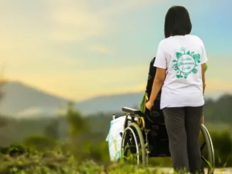 A woman in a white t-shirt stands behind an elderly person in a wheelchair, both facing away from the camera toward a soft, hazy mountain landscape at sunset.