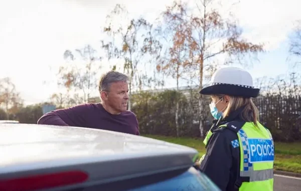 A middle-aged man leaning on his car while calmly speaking to a female police officer wearing a high-visibility vest and a face mask outdoors.