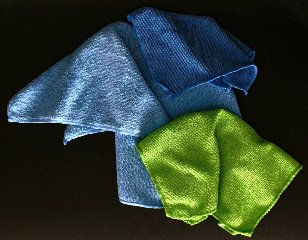 Three folded microfiber cleaning cloths in shades of light blue, royal blue, and lime green arranged on a dark surface.