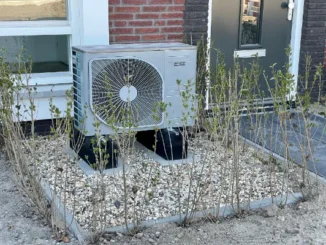 A modern silver air-source heat pump outdoor unit installed on a gravel bed with black support feet in front of a brick house.