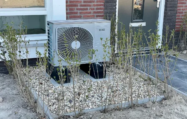 A modern silver air-source heat pump outdoor unit installed on a gravel bed with black support feet in front of a brick house.