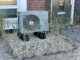 A modern silver air-source heat pump outdoor unit installed on a gravel bed with black support feet in front of a brick house.