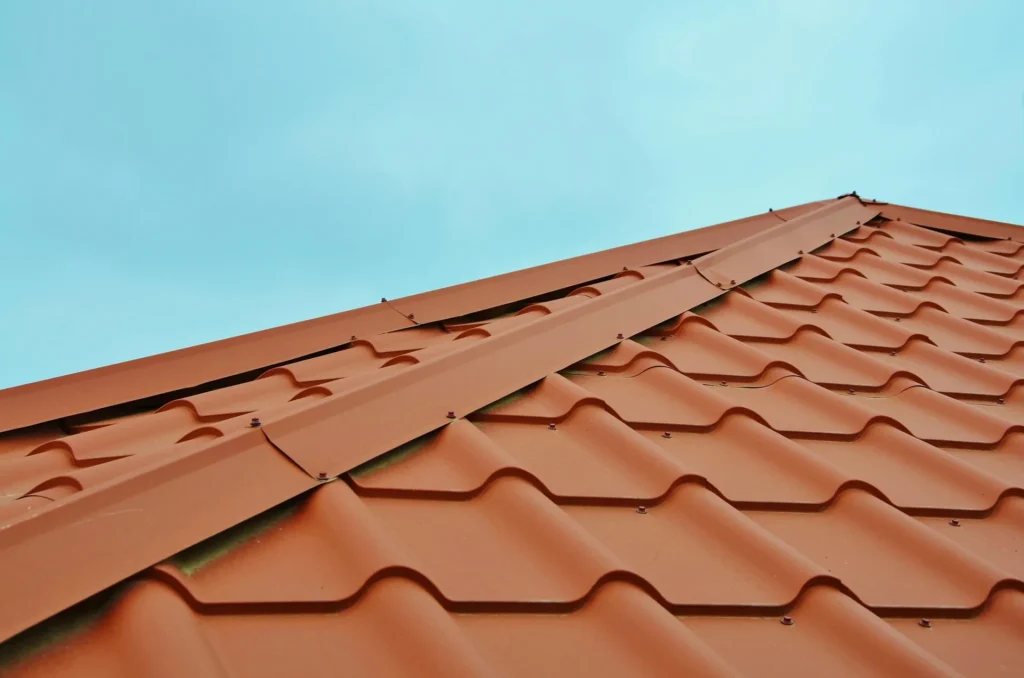 A close-up, low-angle shot of a new terracotta-colored metal tile roof with a clean ridge cap installation against a clear blue sky.
