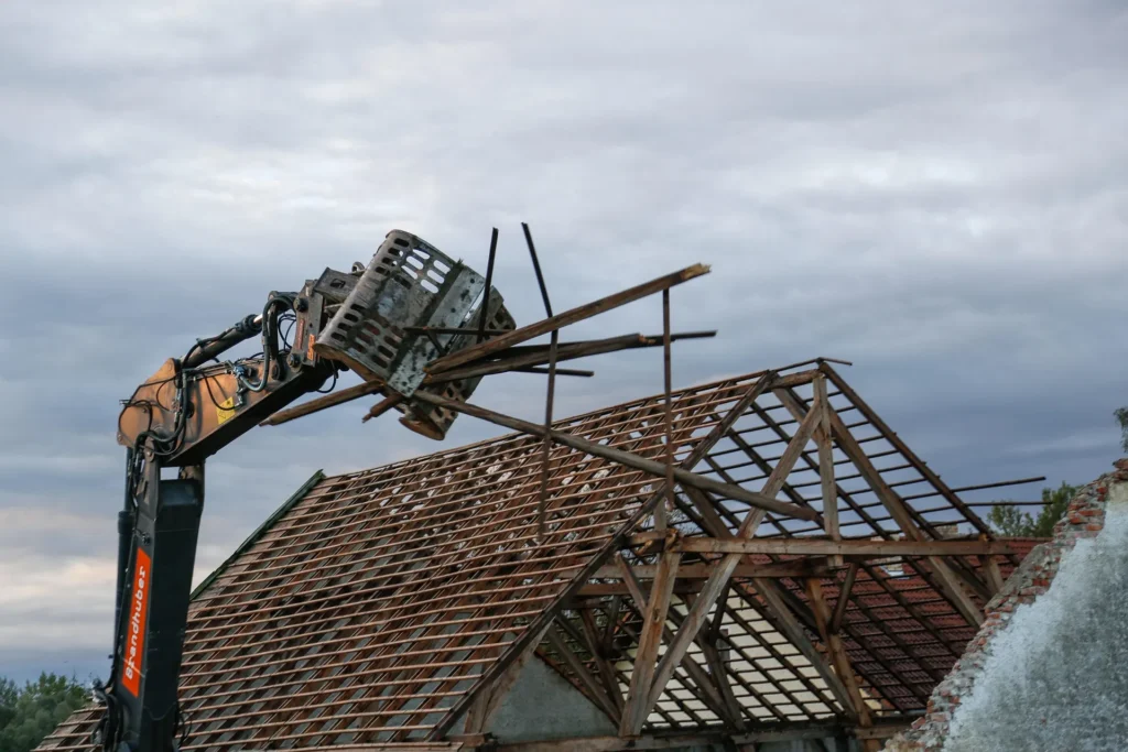 A hydraulic excavator arm lifting wooden beams over the exposed timber frame of a roof under construction.