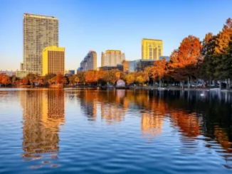 A scenic view of the downtown Orlando skyline reflecting off Lake Eola at sunset, featuring the tall residential VUE building and orange-leafed cypress trees along the shore.