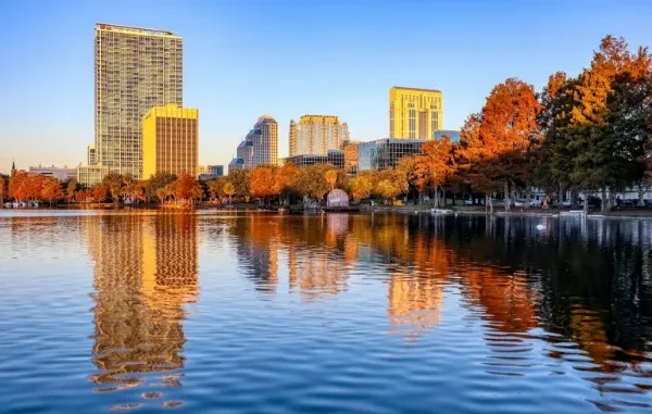 A scenic view of the downtown Orlando skyline reflecting off Lake Eola at sunset, featuring the tall residential VUE building and orange-leafed cypress trees along the shore.