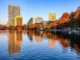 A scenic view of the downtown Orlando skyline reflecting off Lake Eola at sunset, featuring the tall residential VUE building and orange-leafed cypress trees along the shore.