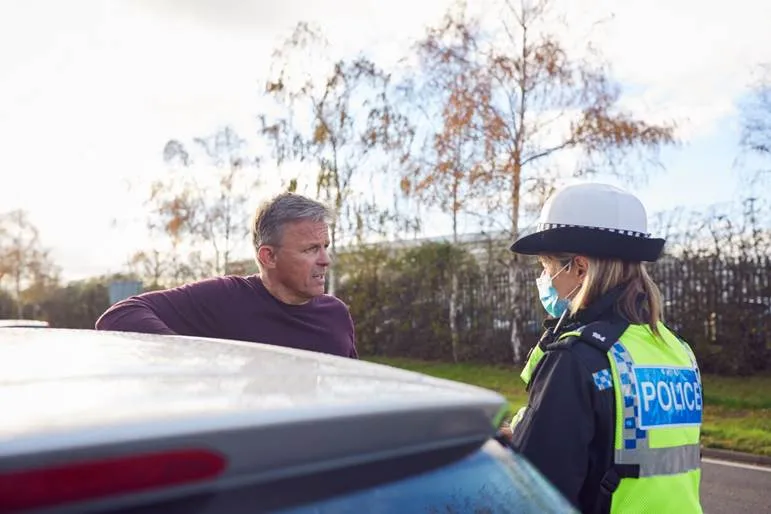 A middle-aged man stands next to his car, speaking calmly with a female police officer wearing a high-visibility vest and a face mask during a daytime roadside stop.