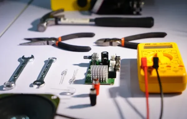 A collection of professional tools including a yellow digital multimeter, circuit board, needle-nose pliers, wire cutters, wrenches, and a hammer arranged on a white workbench.