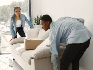 A man and a woman in denim shirts working together to carefully lift and position a light-colored sofa in a bright living room during a move.