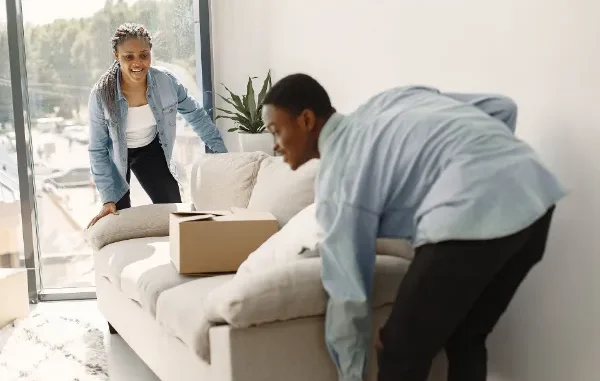 A man and a woman in denim shirts working together to carefully lift and position a light-colored sofa in a bright living room during a move.