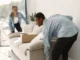 A man and a woman in denim shirts working together to carefully lift and position a light-colored sofa in a bright living room during a move.