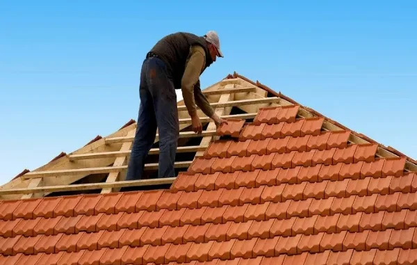A professional roofer carefully installing terracotta clay tiles on a wooden roof frame against a clear blue sky.
