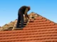 A professional roofer carefully installing terracotta clay tiles on a wooden roof frame against a clear blue sky.