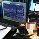 A professional trading desk featuring a Samsung laptop, tablet, and smartphone displaying GameStop (GME) stock charts and financial data alongside cash and a notebook.