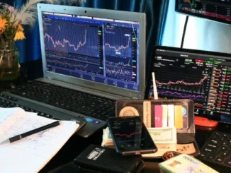 A professional trading desk featuring a Samsung laptop, tablet, and smartphone displaying GameStop (GME) stock charts and financial data alongside cash and a notebook.