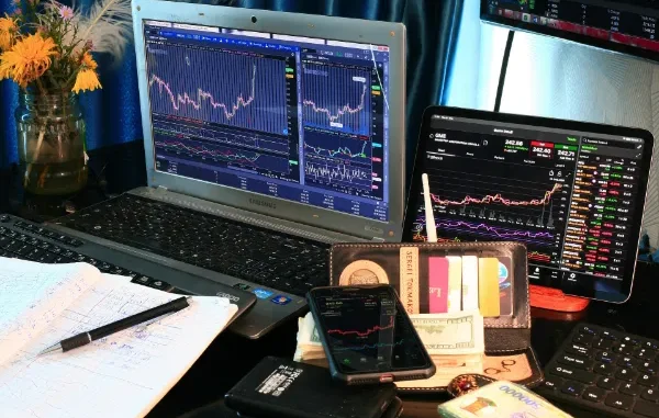 A professional trading desk featuring a Samsung laptop, tablet, and smartphone displaying GameStop (GME) stock charts and financial data alongside cash and a notebook.