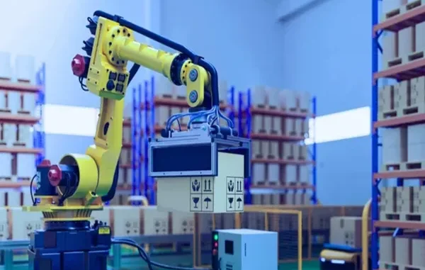 A yellow industrial robotic arm lifting a cardboard box in a high-tech warehouse with rows of shelving in the background.