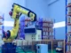 A yellow industrial robotic arm lifting a cardboard box in a high-tech warehouse with rows of shelving in the background.