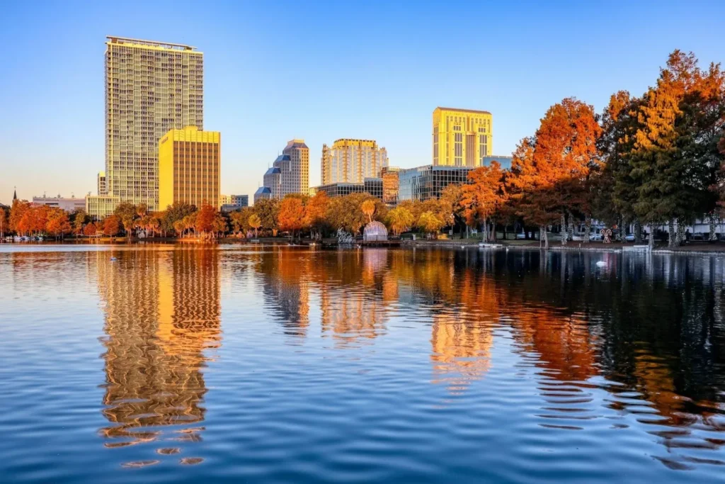 A scenic view of the downtown Orlando skyline reflecting on the calm blue water of Lake Eola at sunset. The image features prominent high-rise buildings, including The VUE at Lake Eola, framed by lush autumn trees with vibrant orange and red leaves.