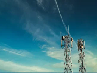 Two tall metal lattice telecommunication towers equipped with various antennas and satellite dishes against a blue sky with thin clouds.