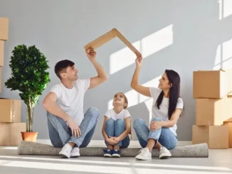 A smiling family of three sitting on a rolled-up rug in a new house, with the parents holding a piece of cardboard over their daughter's head like a roof.