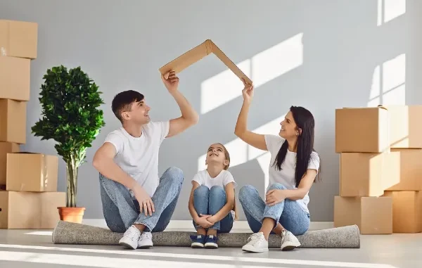 A smiling family of three sitting on a rolled-up rug in a new house, with the parents holding a piece of cardboard over their daughter's head like a roof.