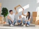 A smiling family of three sitting on a rolled-up rug in a new house, with the parents holding a piece of cardboard over their daughter's head like a roof.