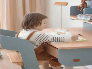 A young child sitting in a blue and natural wood Boori ergonomic chair, reading a book at a matching Boori study desk in a brightly lit bedroom.