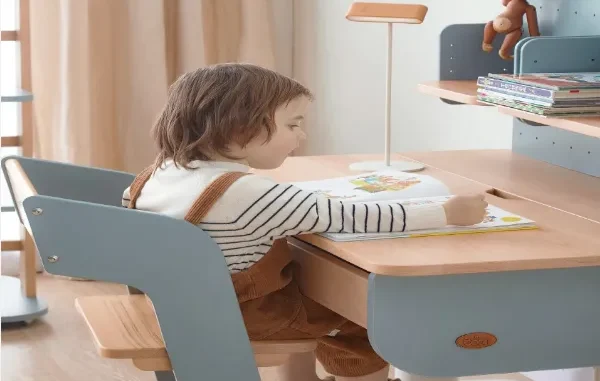 A young child sitting in a blue and natural wood Boori ergonomic chair, reading a book at a matching Boori study desk in a brightly lit bedroom.